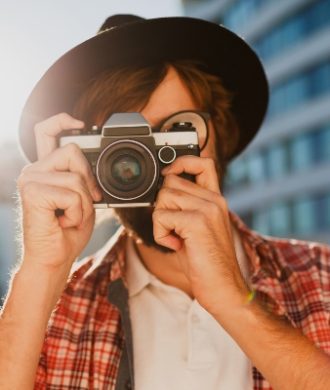 Fashionable  beard man portrait, using retro film photo camera  , making  photos .  Hipster style. Wearing black hat, stylish eyeglasses, checkered shirt .